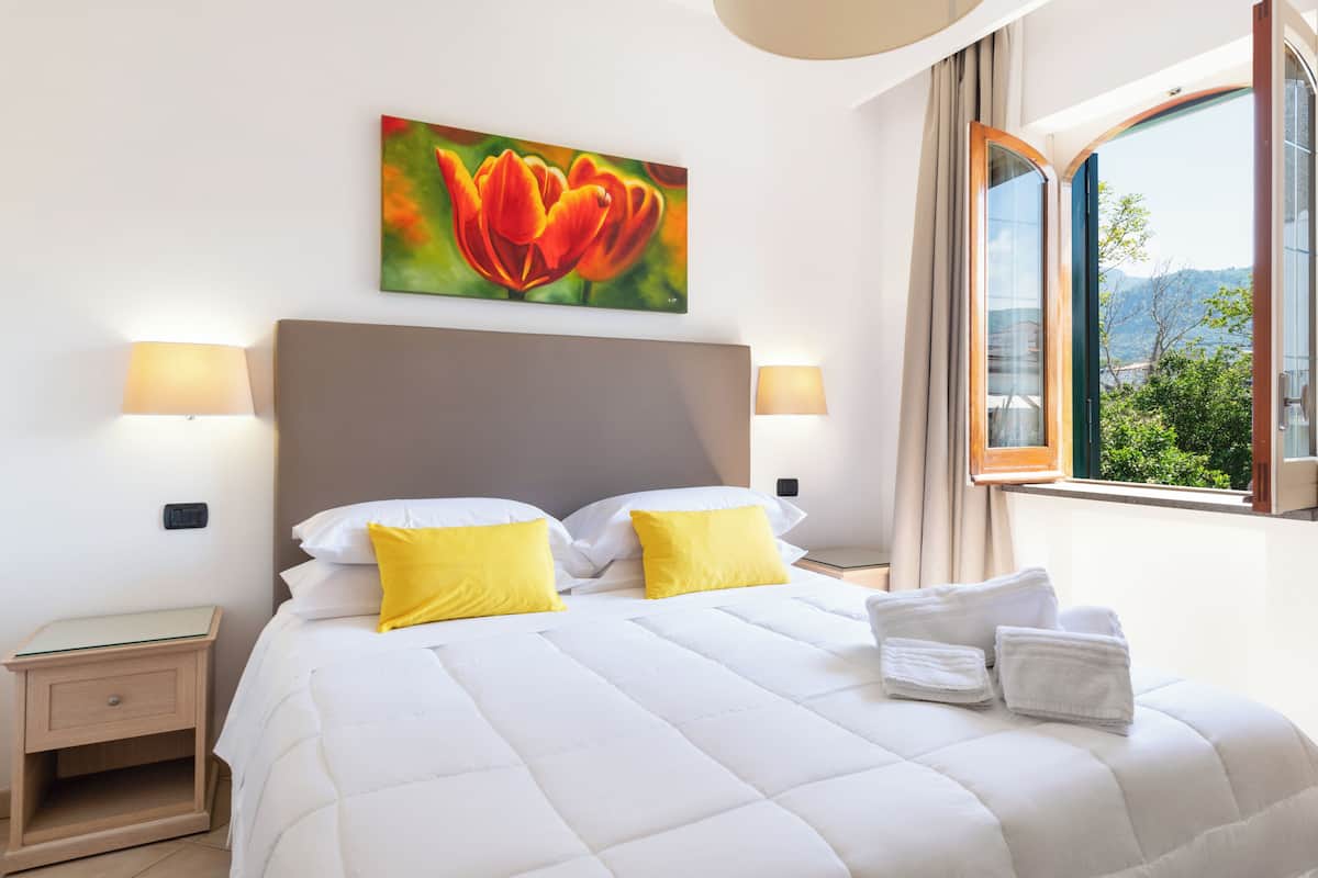 A hotel room with a large bed, two bedside lamps, a painting of a flower, and a window with a view of greenery and mountains.