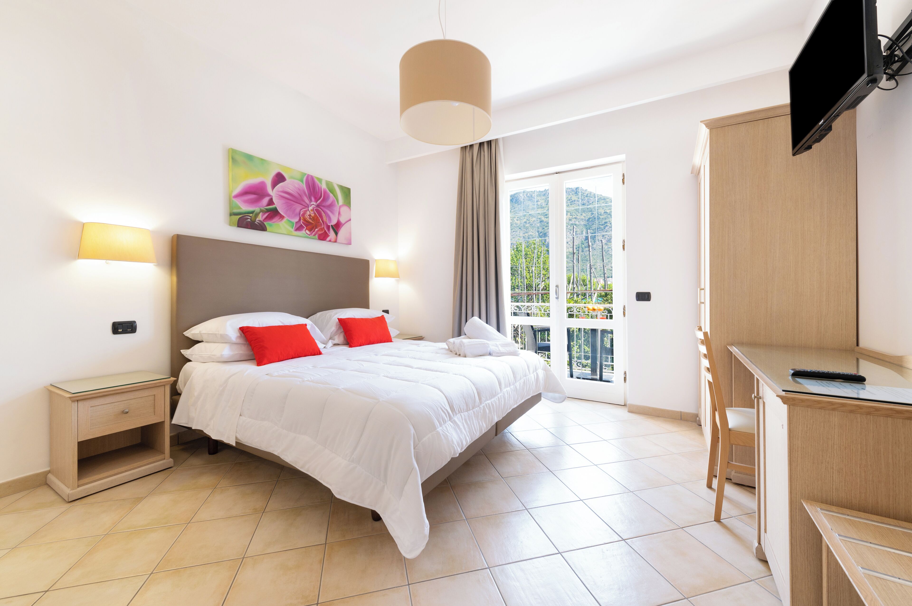 A bedroom with a bed, bedside tables, a desk, a chair, a TV, and a balcony with a view.