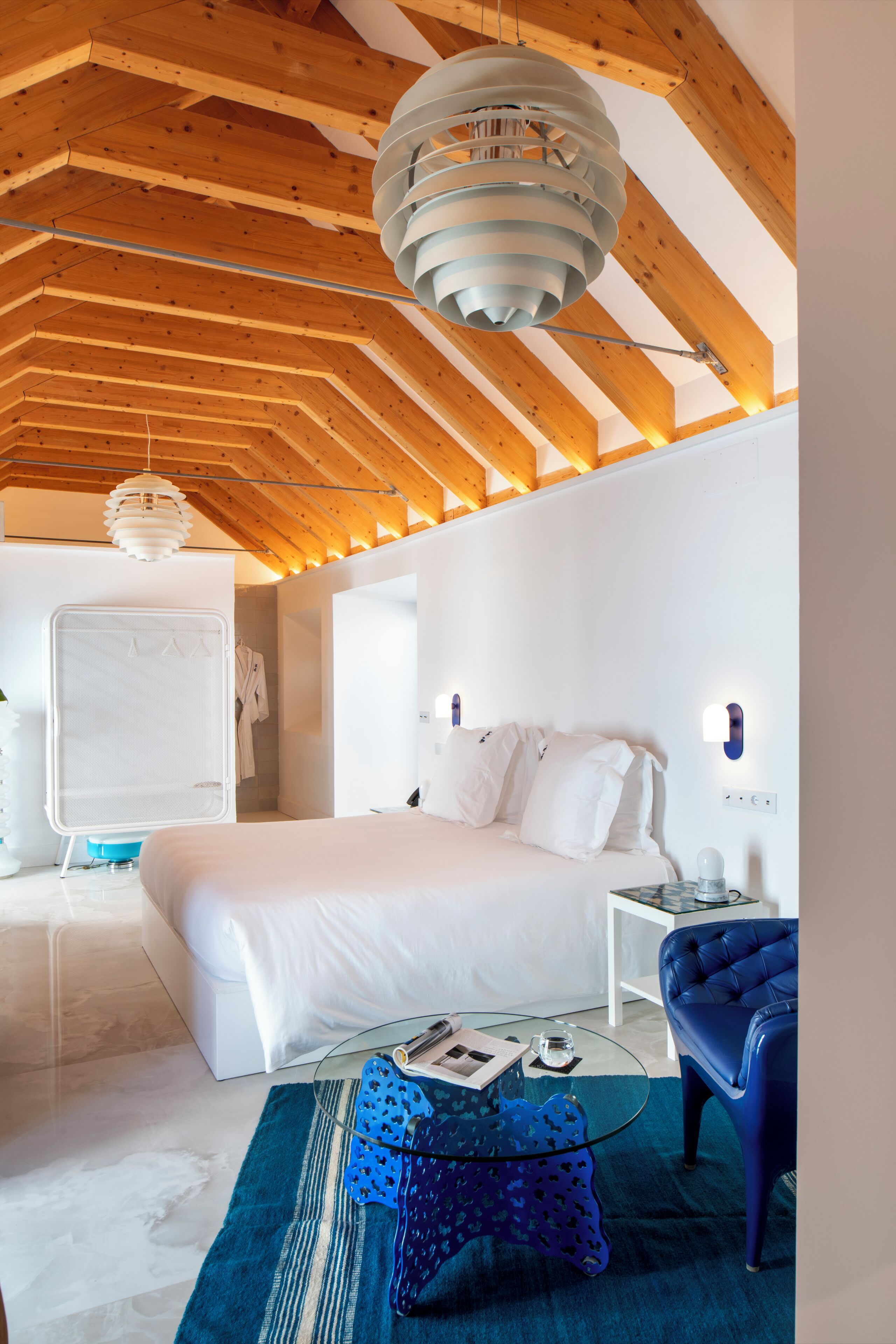 A modern bedroom with a large bed, a glass coffee table, a blue chair, and a unique ceiling light fixture.