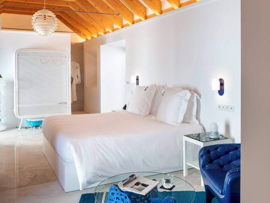 A modern bedroom with a large bed, a glass coffee table, a blue chair, and a unique ceiling light fixture.
