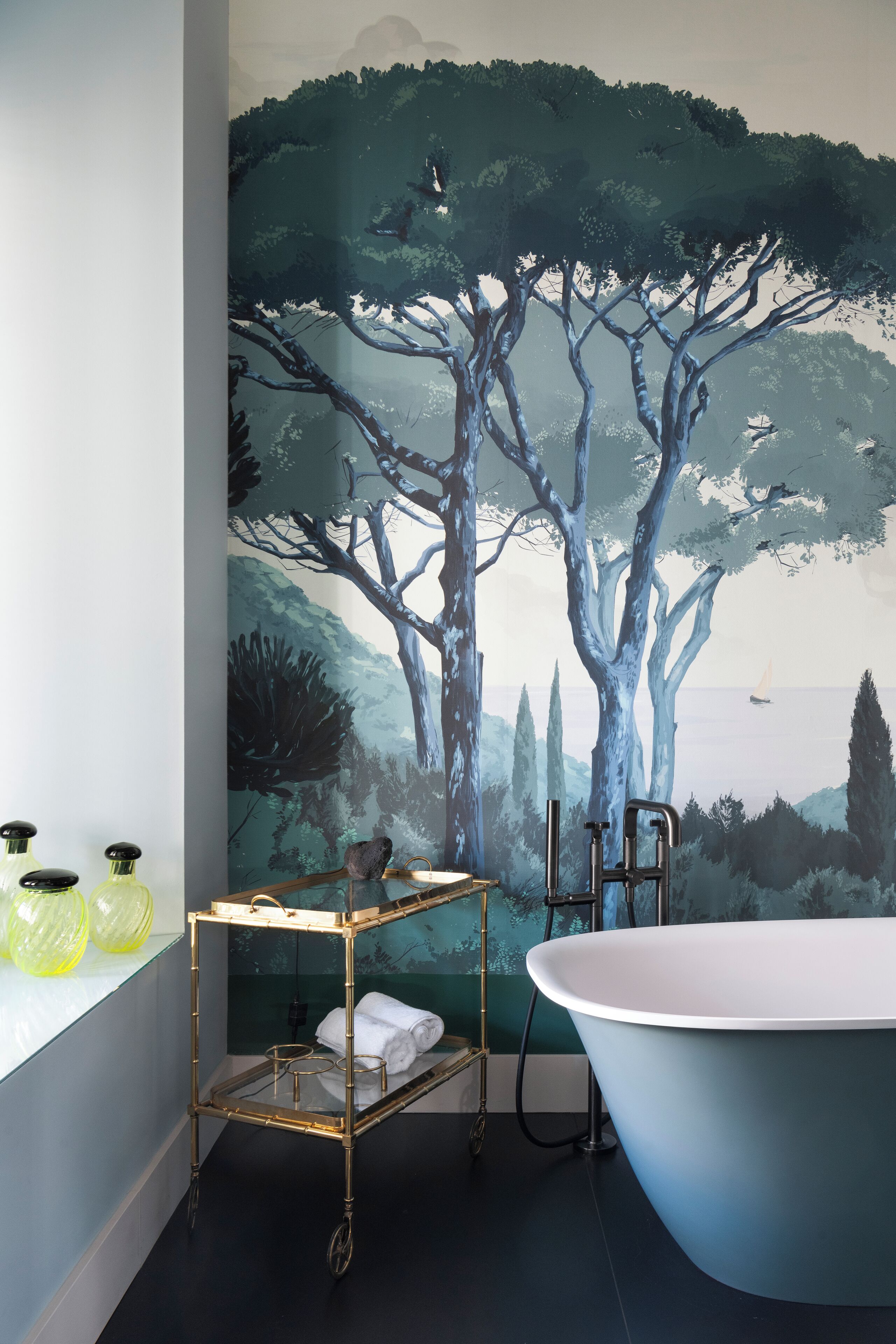 A bathroom with a large bathtub, a gold cart with toiletries, and a mural of trees and a landscape.