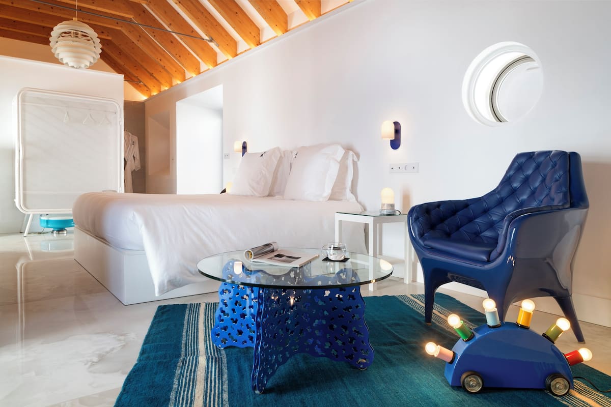 A modern bedroom with a bed, a glass coffee table, a blue armchair, and a unique blue table with lights.