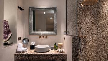 A modern bathroom with a granite countertop, a round sink, a mirror, a towel rack, a small round mirror, a soap dispenser, and a shower with a granite wall.