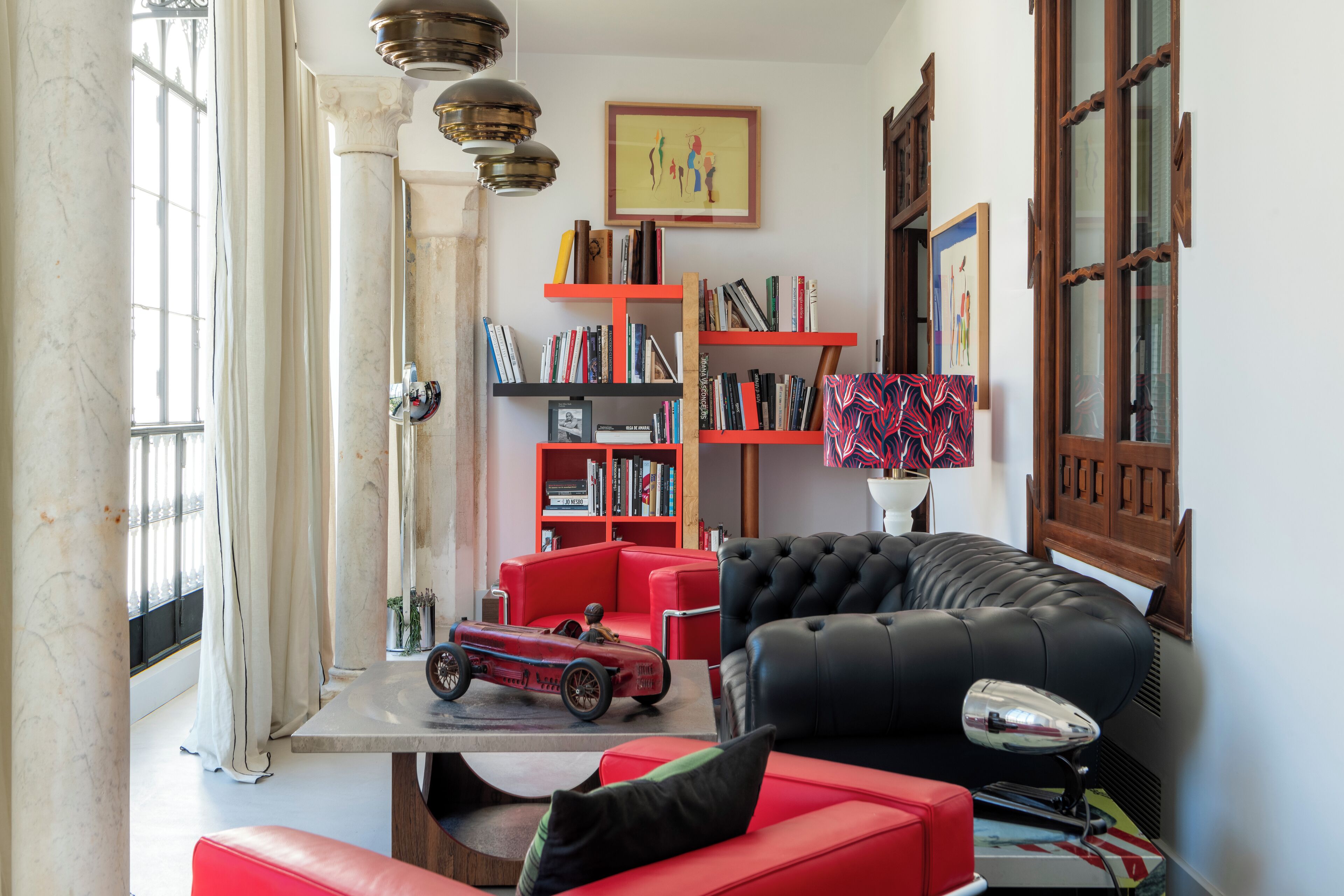 A living room with a red sofa, a black leather couch, and a red armchair. There is a bookshelf with books and a small red car model on a table.