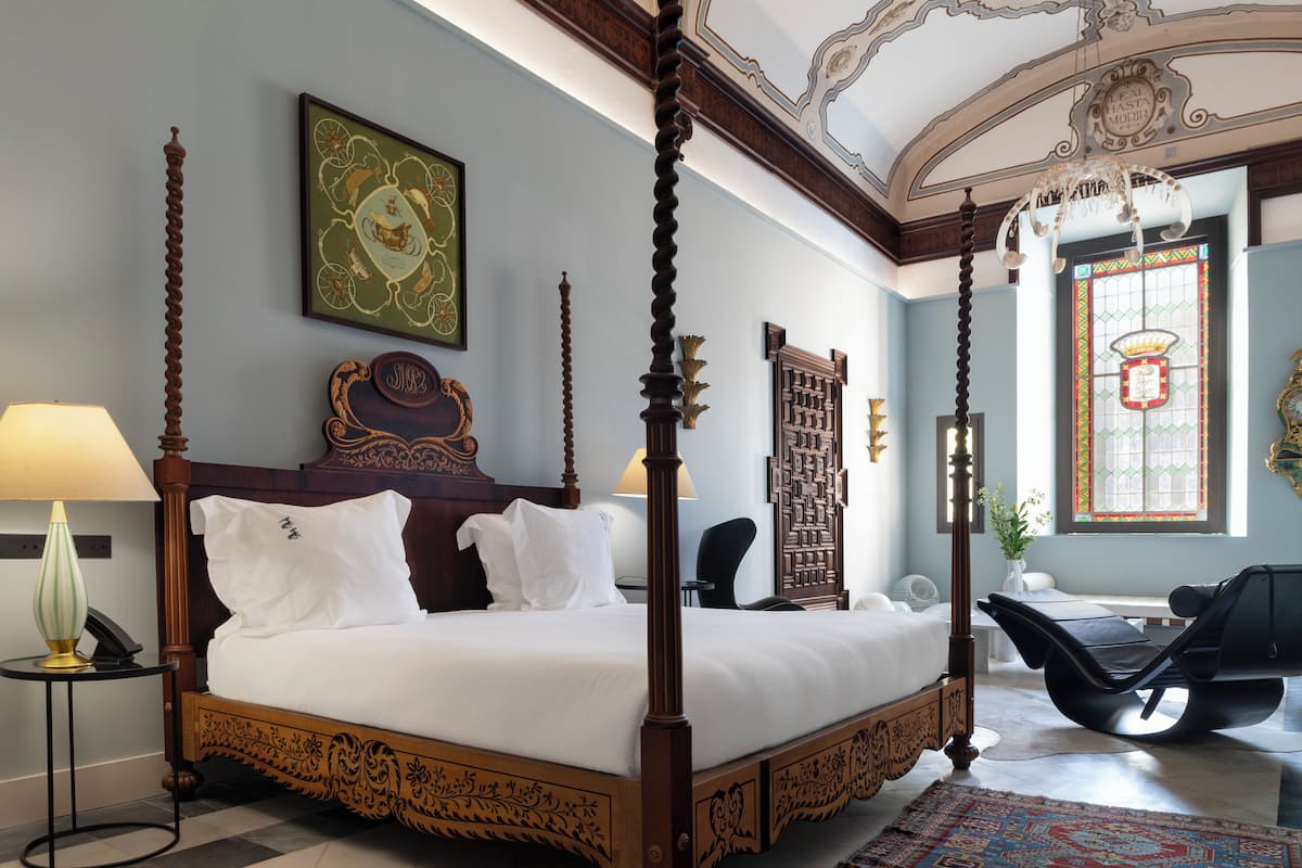 A four-poster bed with a wooden frame and white bedding, a small round table with a lamp, a chair, and a large window with stained glass.