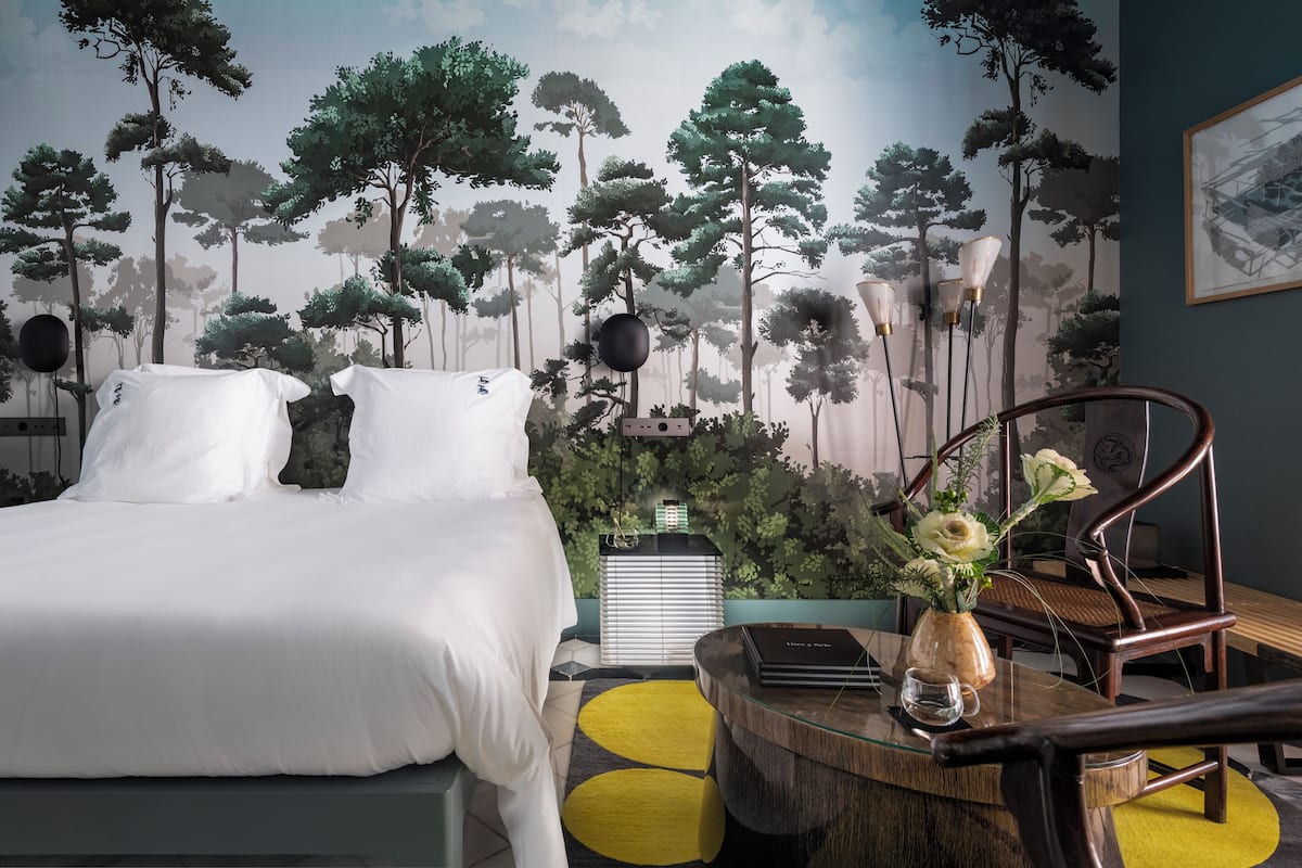 A bedroom with a bed, a nightstand, a chair, a table, and a wall mural of trees.