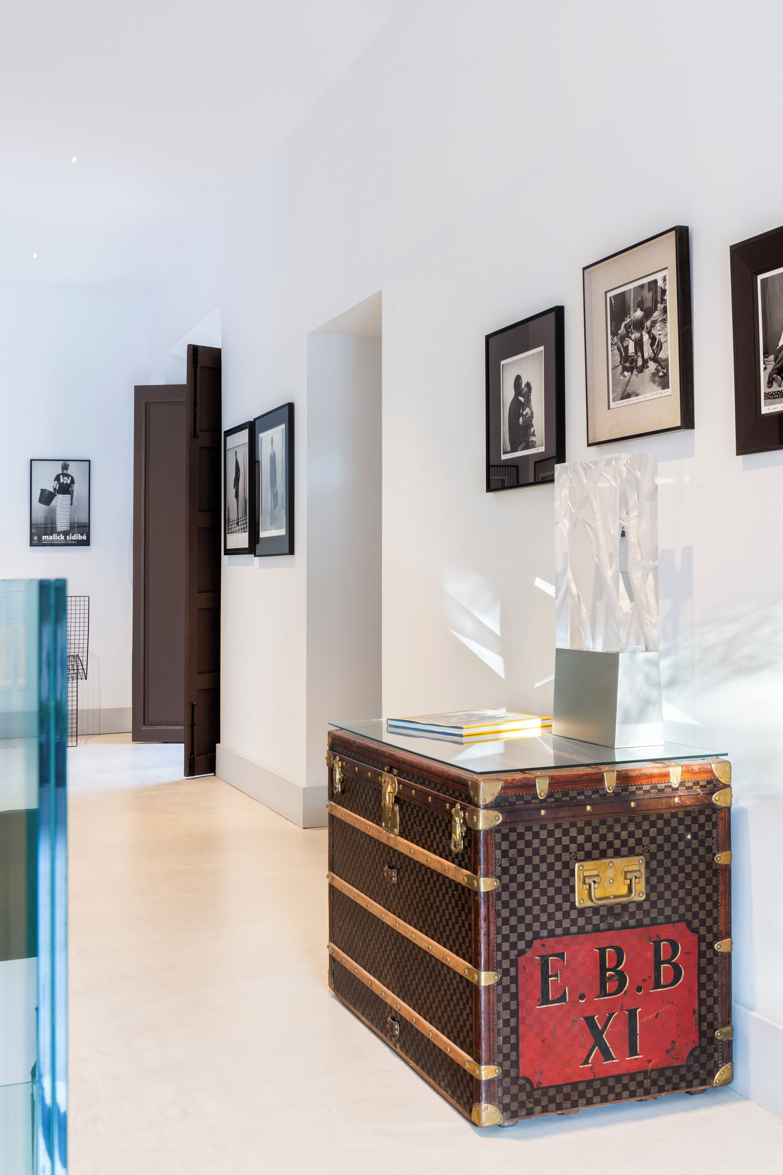 A room with a vintage suitcase labeled 'E.B.B. XI' on a glass table, framed pictures on the wall, and a door leading to another room.