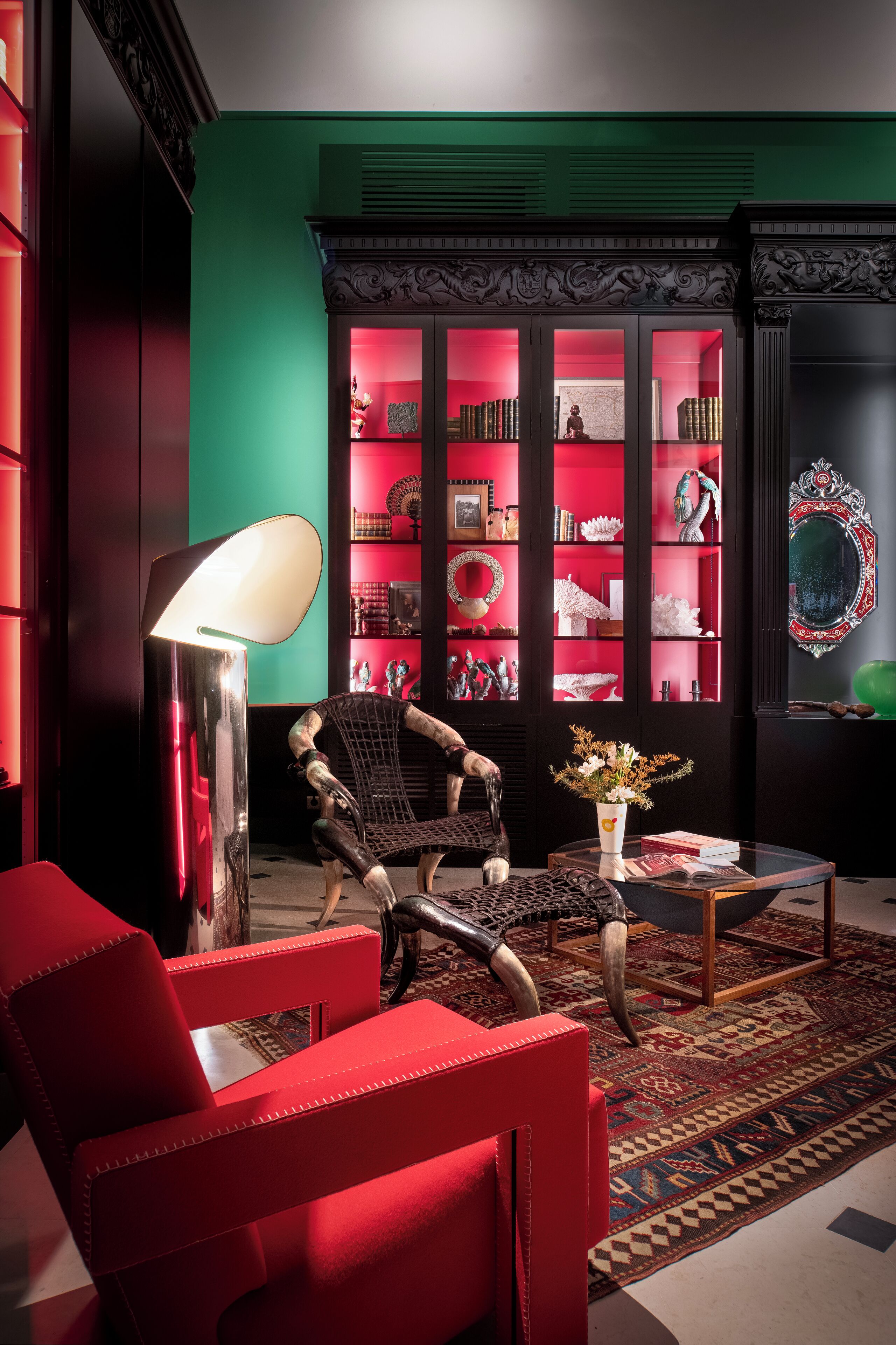 Een moderne woonkamer met een rode bank, een zwarte fauteuil, een vloerlamp en een boekenkast vol decoratieve objecten.