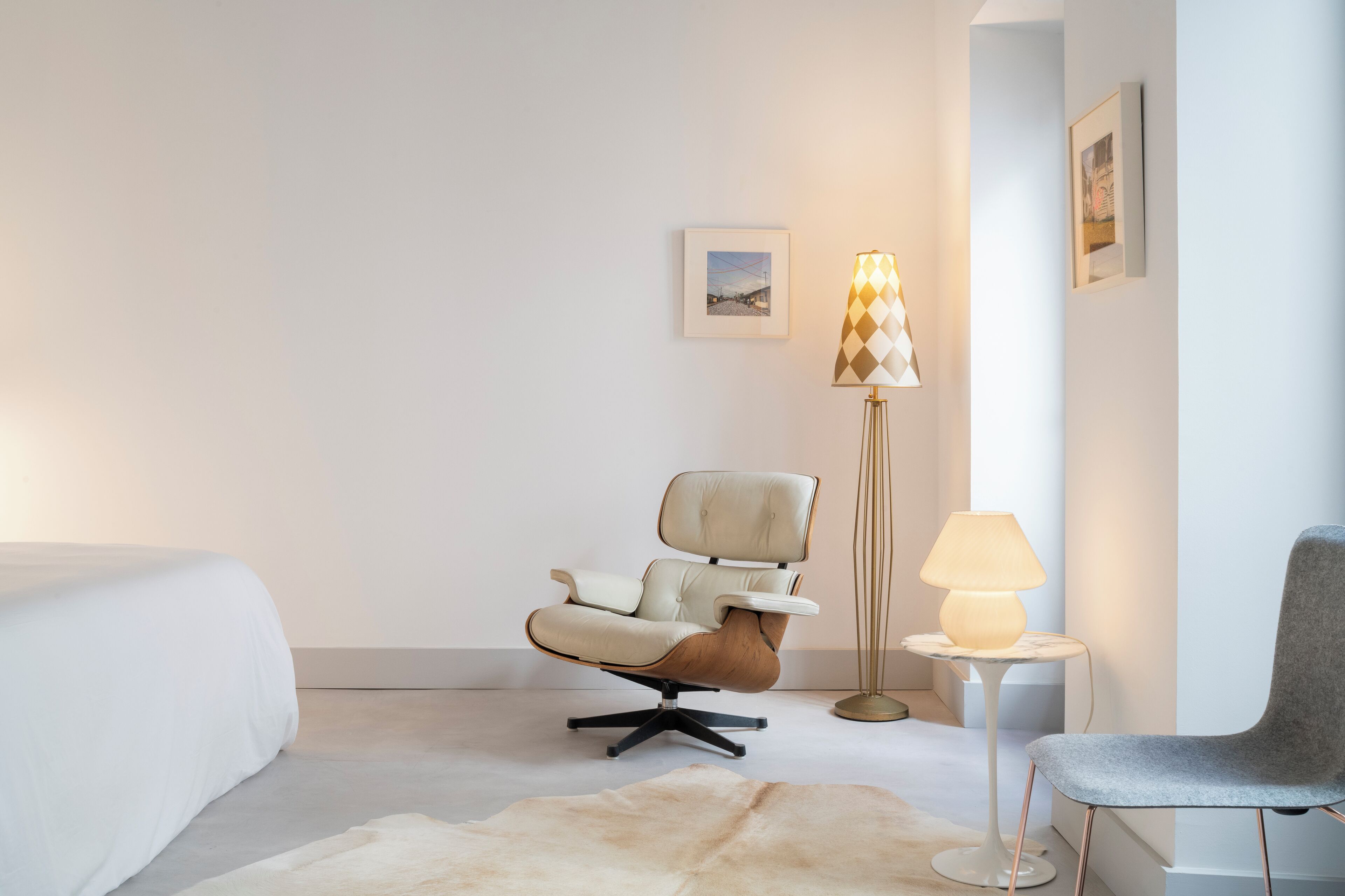 A hotel room with a bed, a modern armchair, a small table with a lamp, and a grey chair.