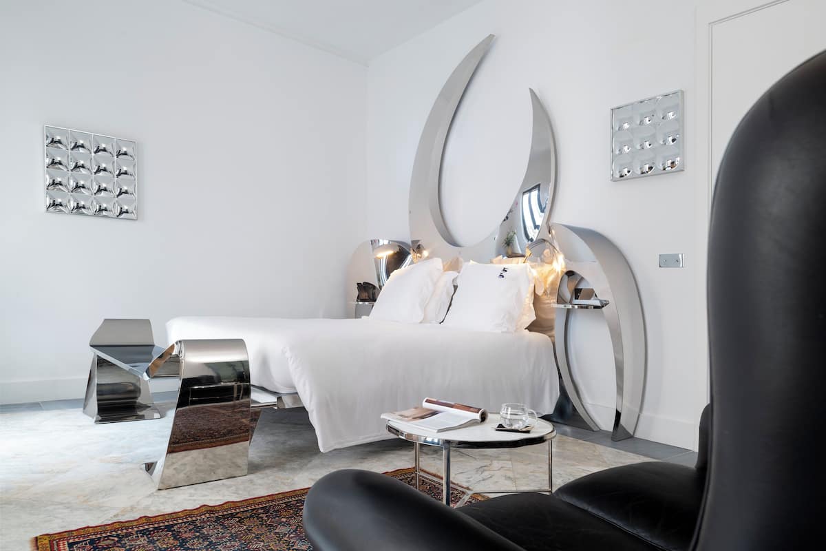 A modern hotel room with a neatly made bed, a small table with a book and glass, and a black chair.