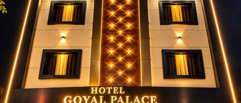 A hotel entrance with a prominent sign reading 'HOTEL GOYAL PALACE' and a modern facade with illuminated windows and a central light fixture.