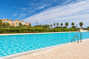 A swimming pool with clear blue water, surrounded by a tiled deck. In the background, there are buildings with multiple balconies and palm trees.