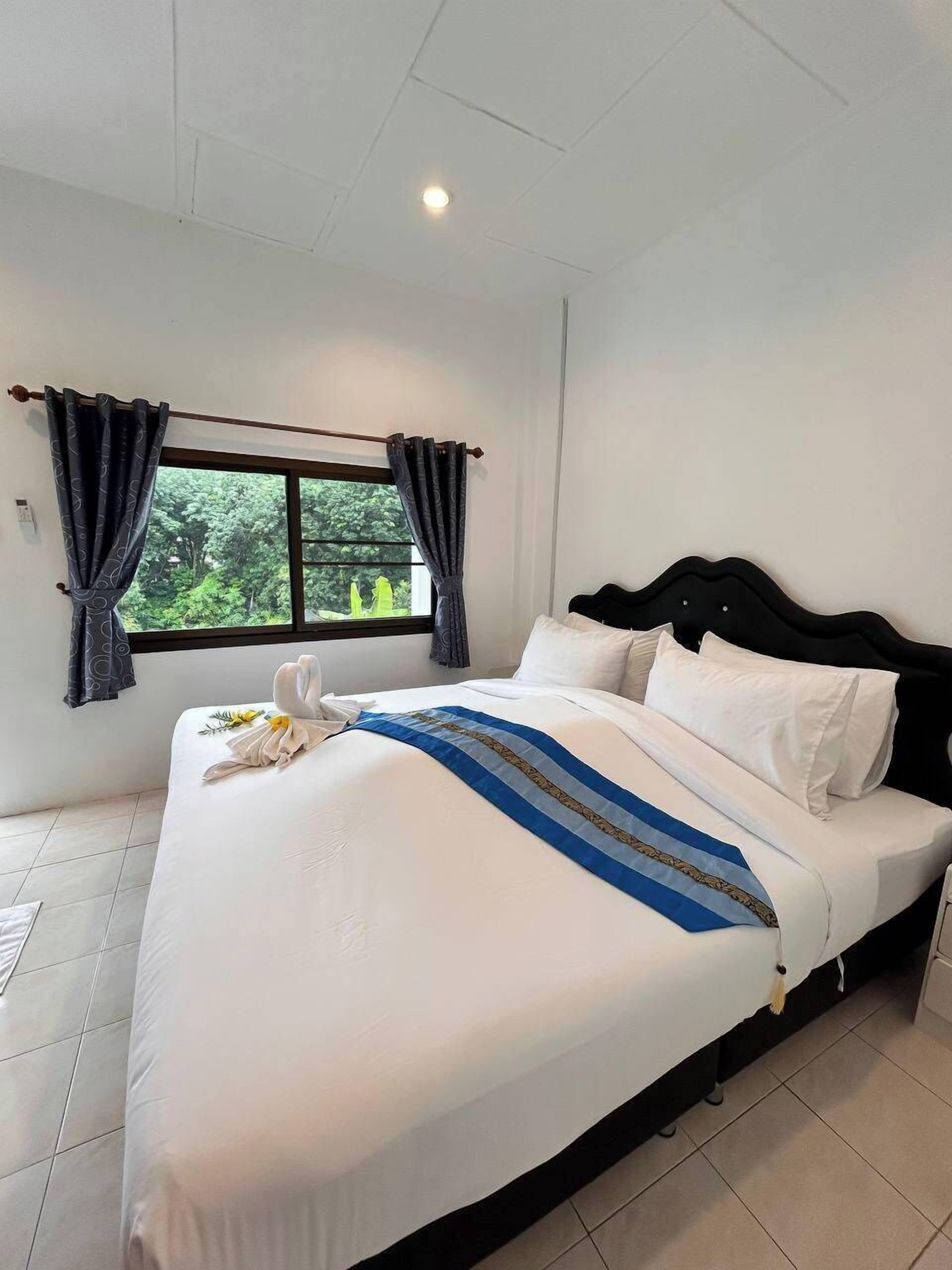 Una cama bien hecha con sábanas blancas y una franja decorativa azul, una ventana con cortinas, y vistas al exterior con vegetación.