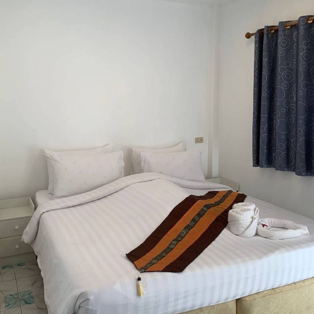 Een netjes opgemaakt bed met witte lakens en een bruin gestreept dekbed, een nachtkastje met een lamp, en een raam met donkere gordijnen.