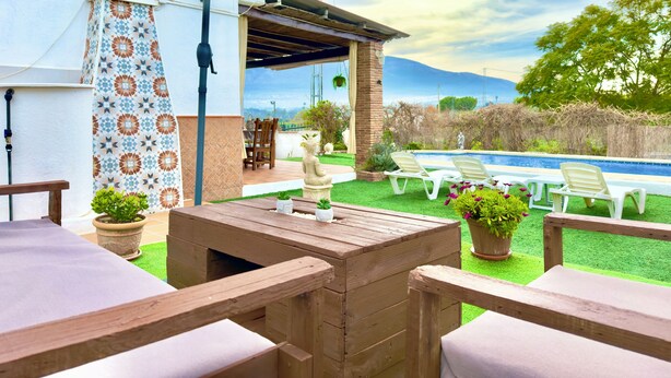 Un patio exterior con una mesa y sillas de madera, un muro con diseño y vistas a una piscina y montañas.