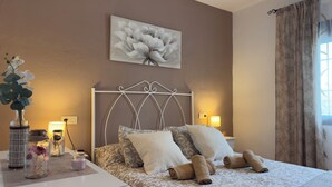 Un dormitorio con cama, almohadas, un florero con flores, una lámpara y un cuadro en la pared.
