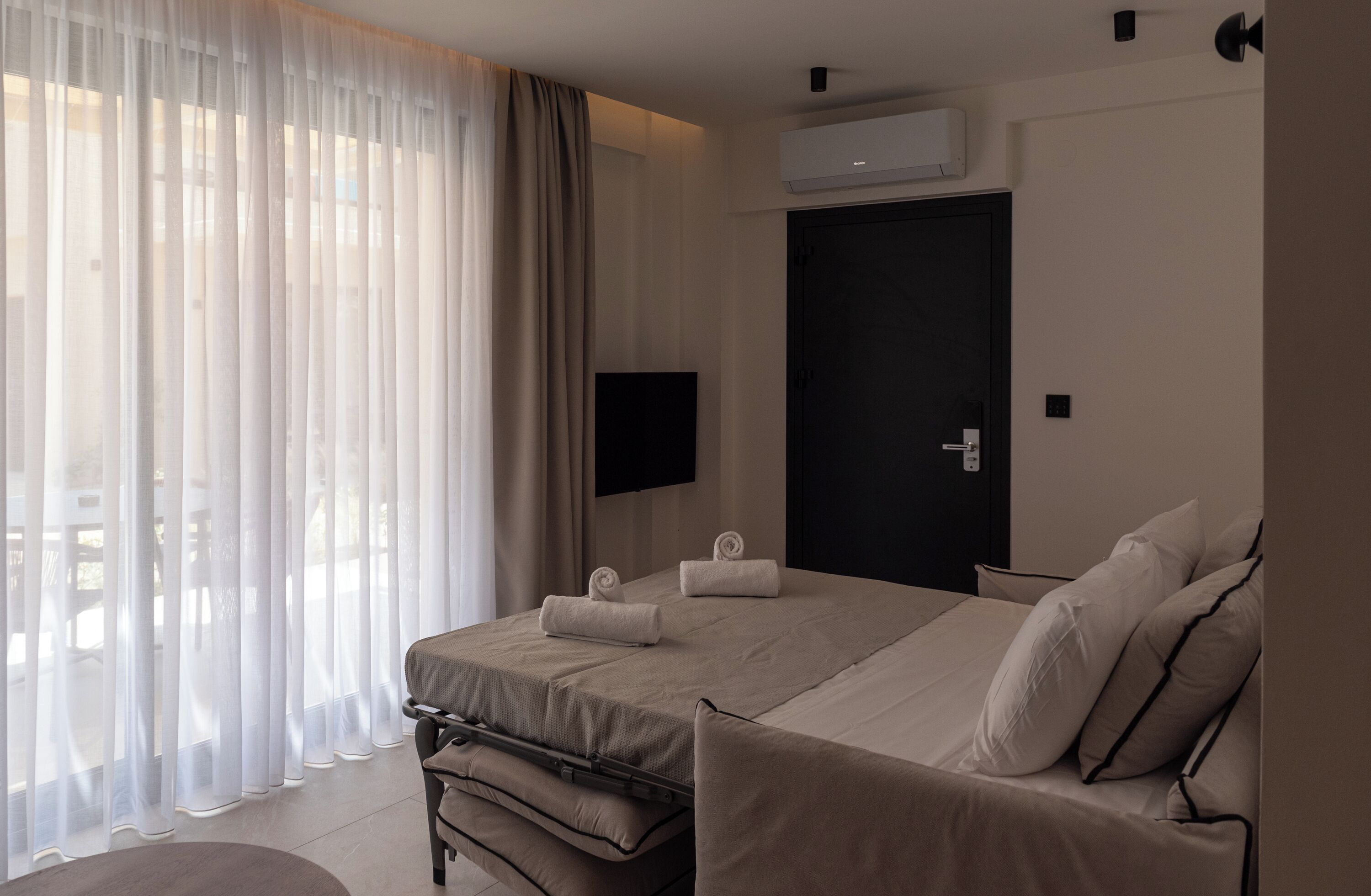 Habitación de hotel moderna con una cama grande, un televisor de pantalla plana, un aire acondicionado y vistas a través de cortinas translúcidas.