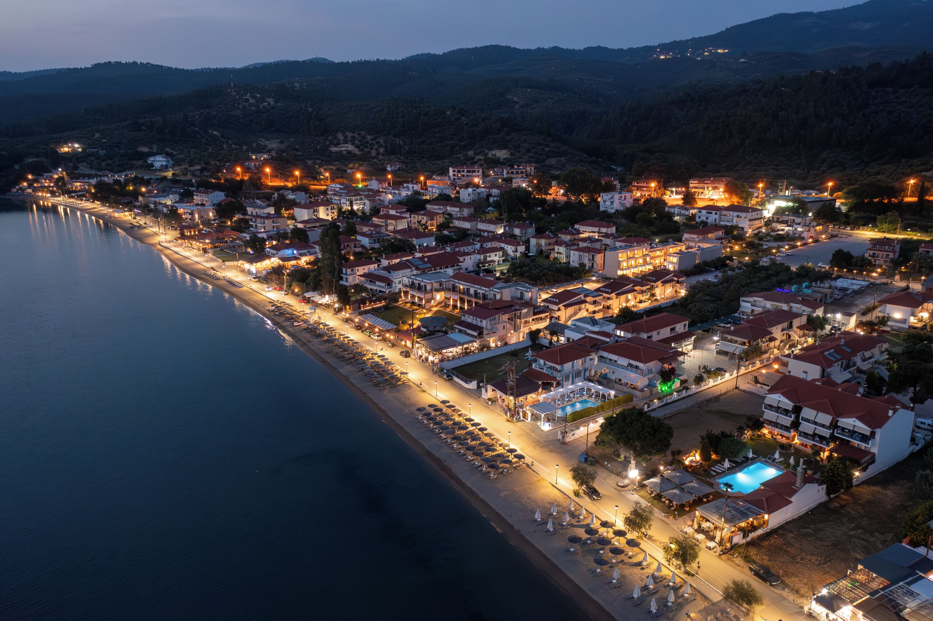 A coastal town at dusk with a beach, hotel buildings, and a swimming pool.
