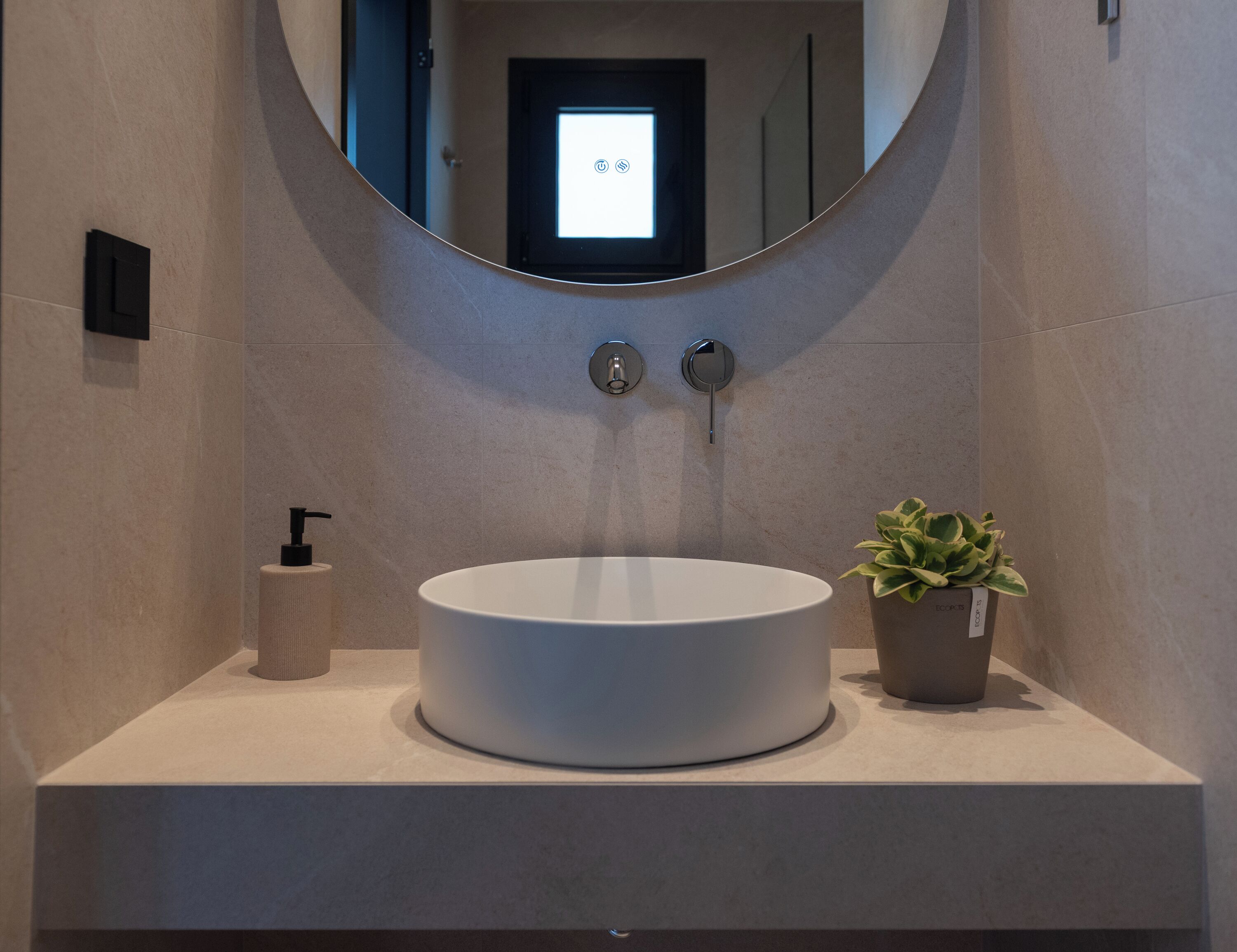 Ein modernes Badezimmer mit einem runden weißen Waschbecken, einer kleinen Topfpflanze und einem Spiegel über dem Waschbecken.