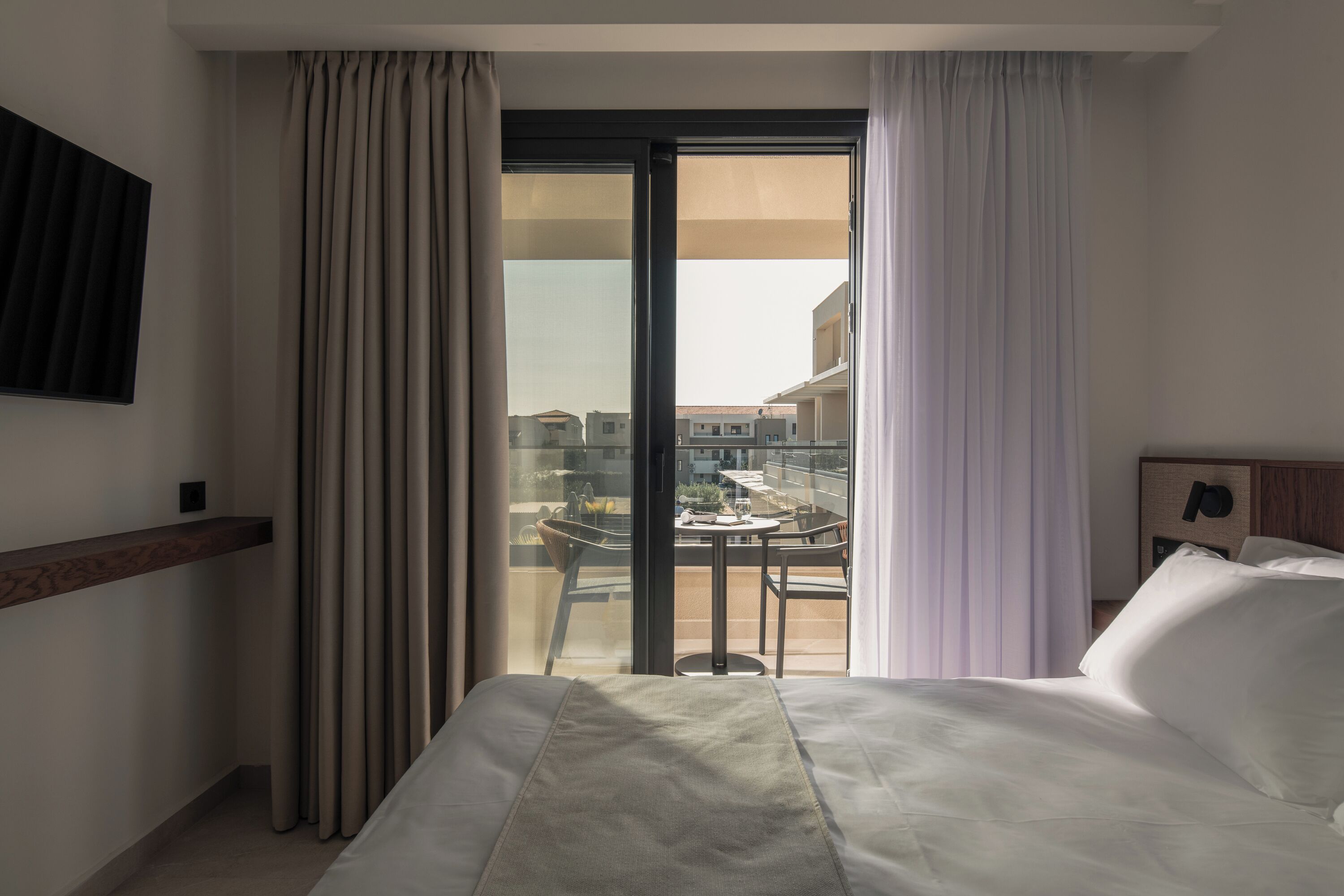 A hotel room with a bed, a view of a balcony with a table and chairs, and a large window with curtains.