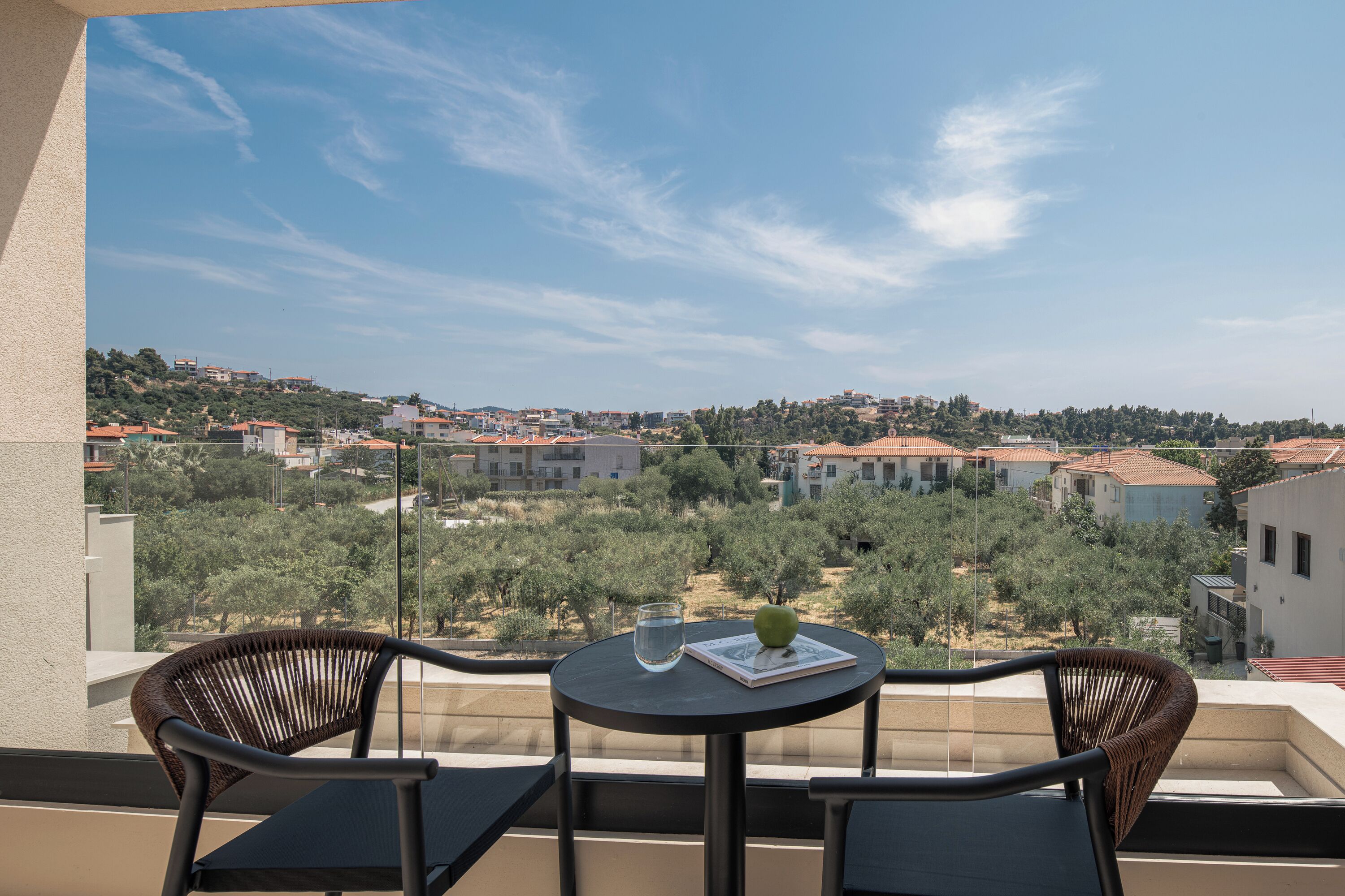 A balcony with wicker chairs and a small round table, overlooking a residential area with trees and buildings.
