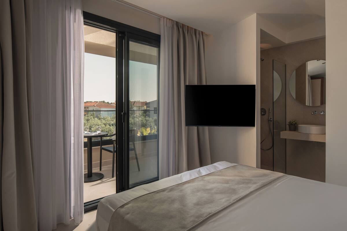 Een hotelkamer met een bed, een aan de muur bevestigde televisie, een badkamer met spiegel en wastafel, en een balkon met een tafel en stoelen.
