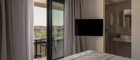 A hotel room with a bed, a TV mounted on the wall, a bathroom with a mirror and sink, and a balcony with a table and chairs.