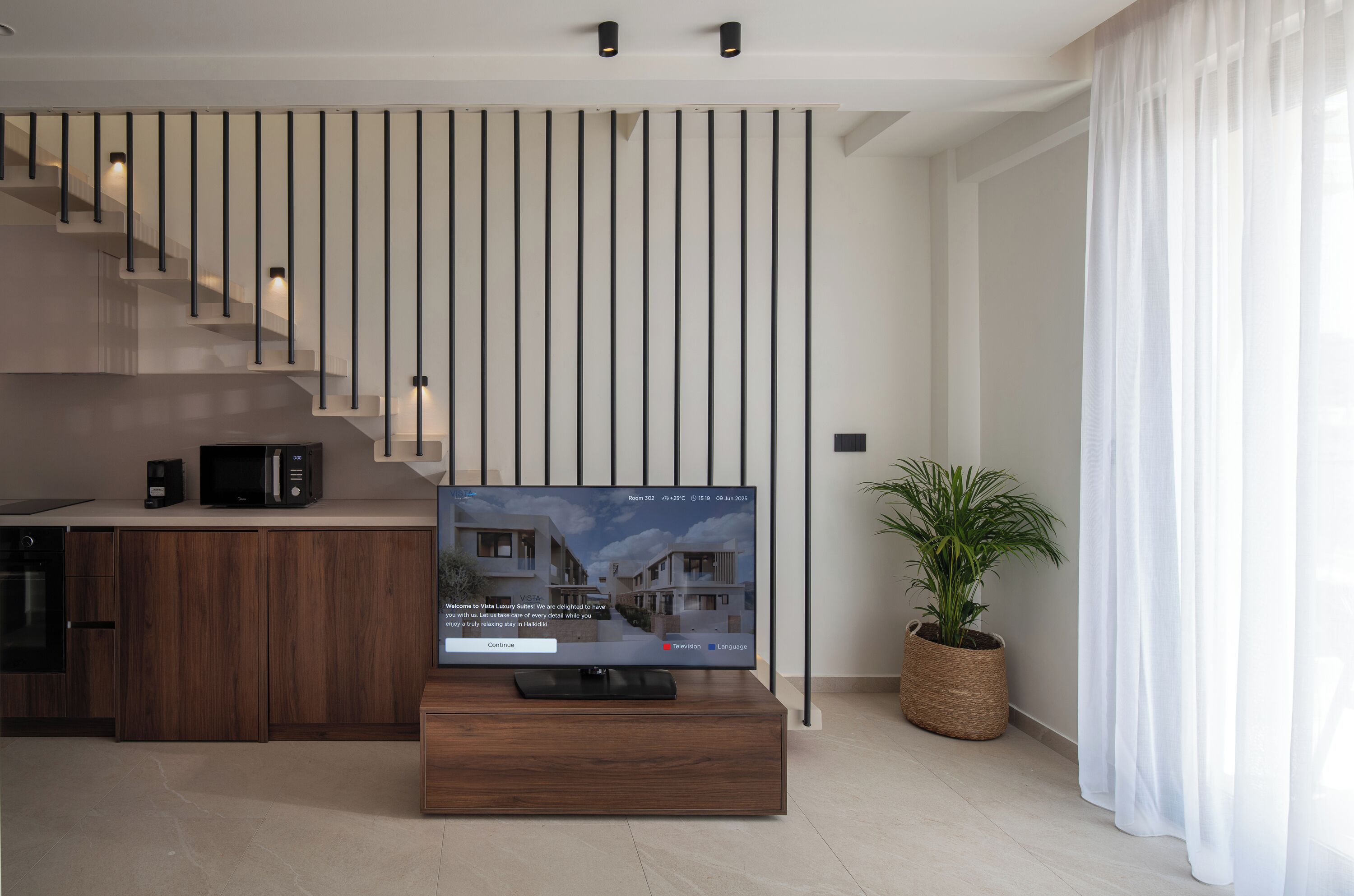 A modern living room with a flat-screen TV on a wooden stand, a kitchen area with a microwave, and a staircase with vertical light fixtures.