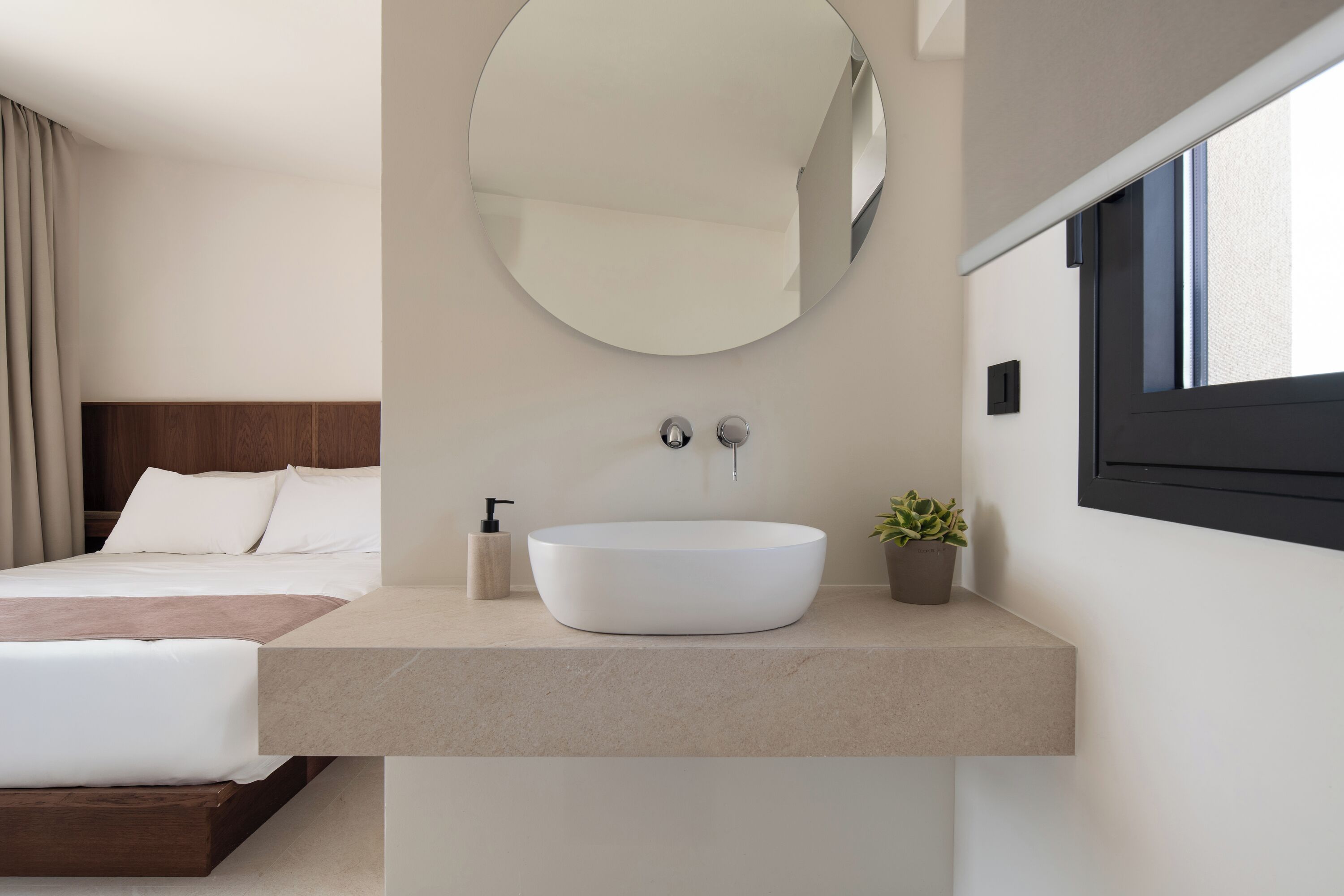 A modern hotel room with a large round mirror, a round white sink, a wooden headboard, a bed with white linens, a small potted plant, and a window with a black frame.