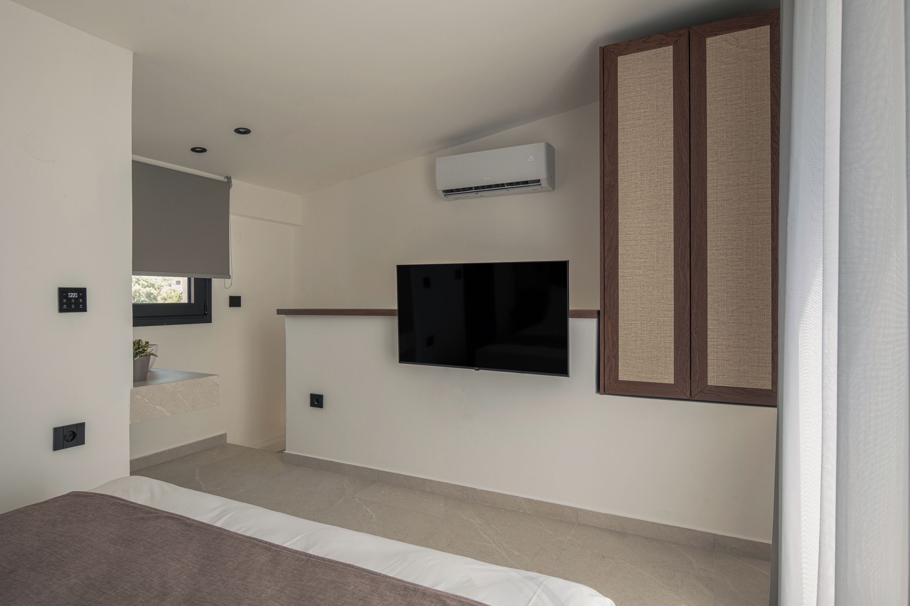 A modern bedroom with a bed, a mounted TV, an air conditioning unit, and a wooden wardrobe.