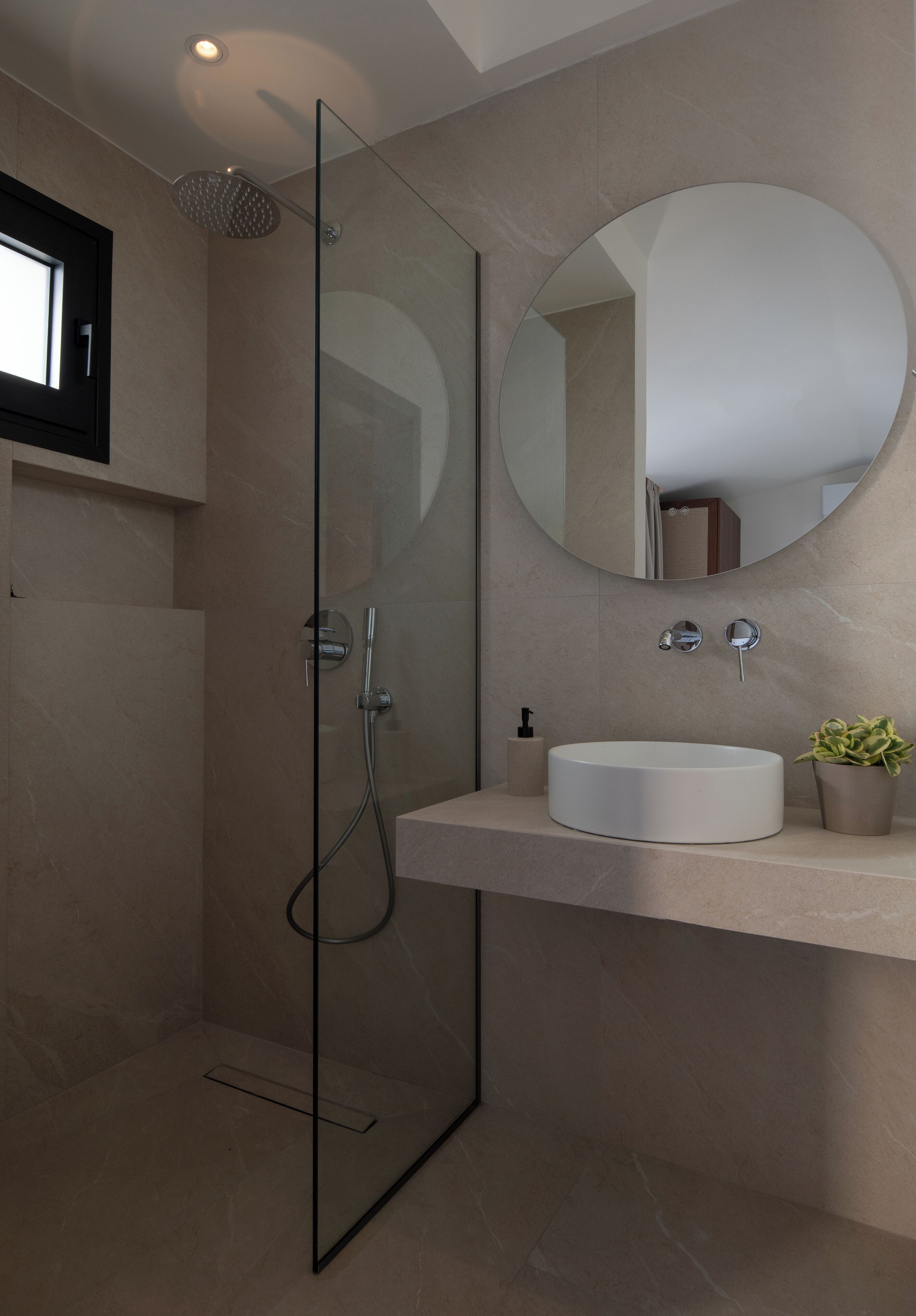 A modern bathroom with a glass-enclosed shower, a round mirror, a white sink, and a small potted plant.