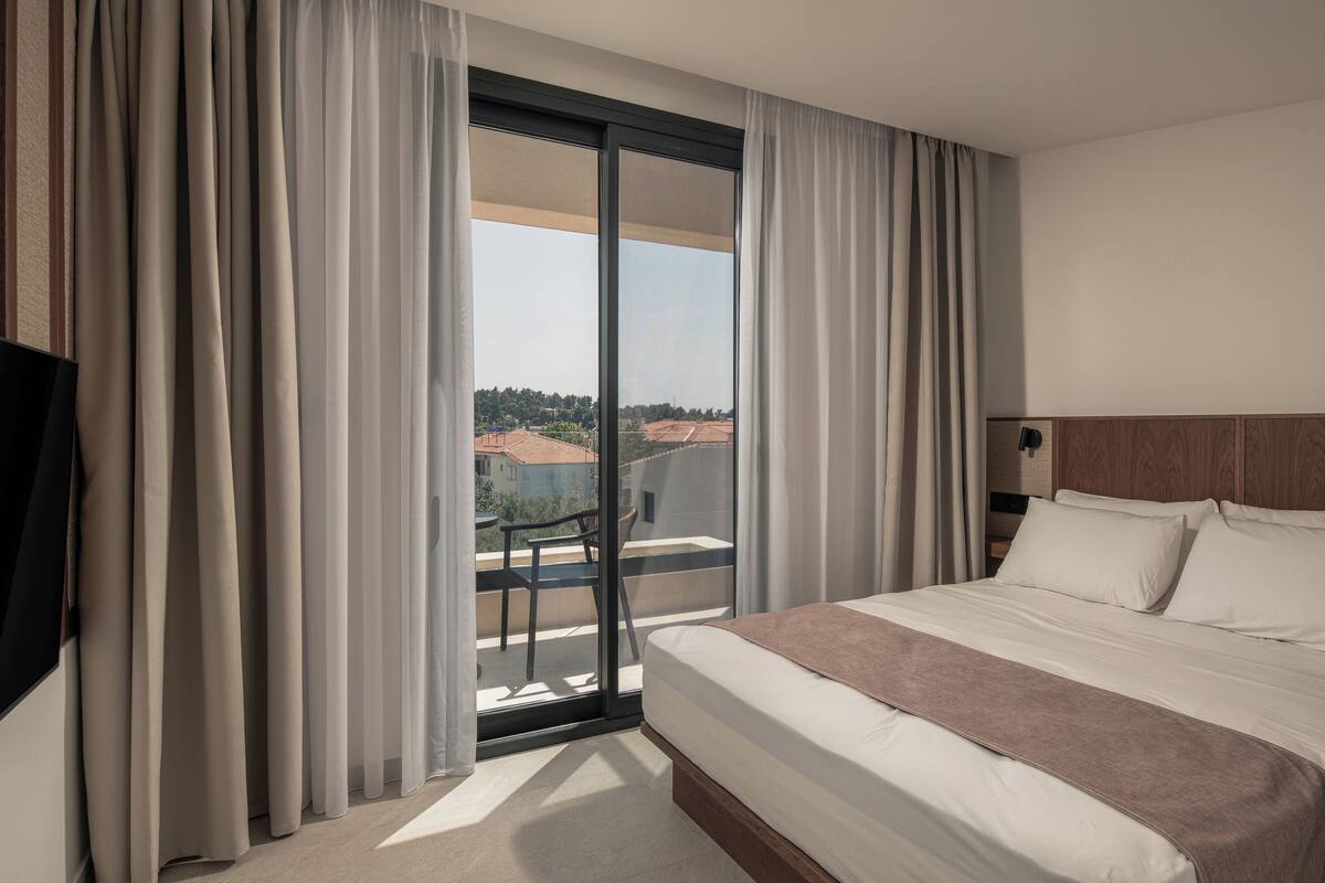 Een hotelkamer met een groot bed, een schuifglazen deur naar een balkon en uitzicht op gebouwen en bomen.