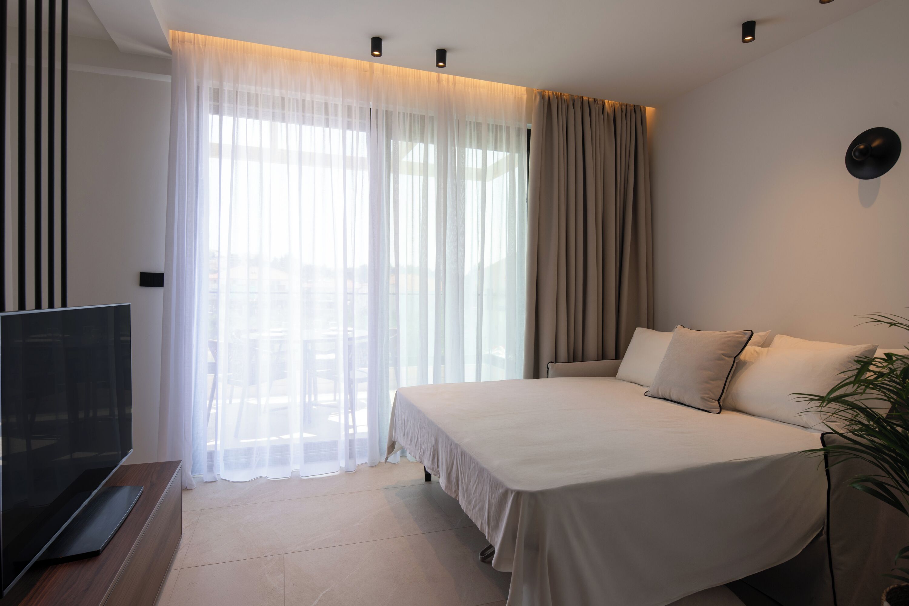 Ein modernes Hotelzimmer mit einem großen Bett, einem Flachbildfernseher und einem großen Fenster mit durchsichtigen Vorhängen.