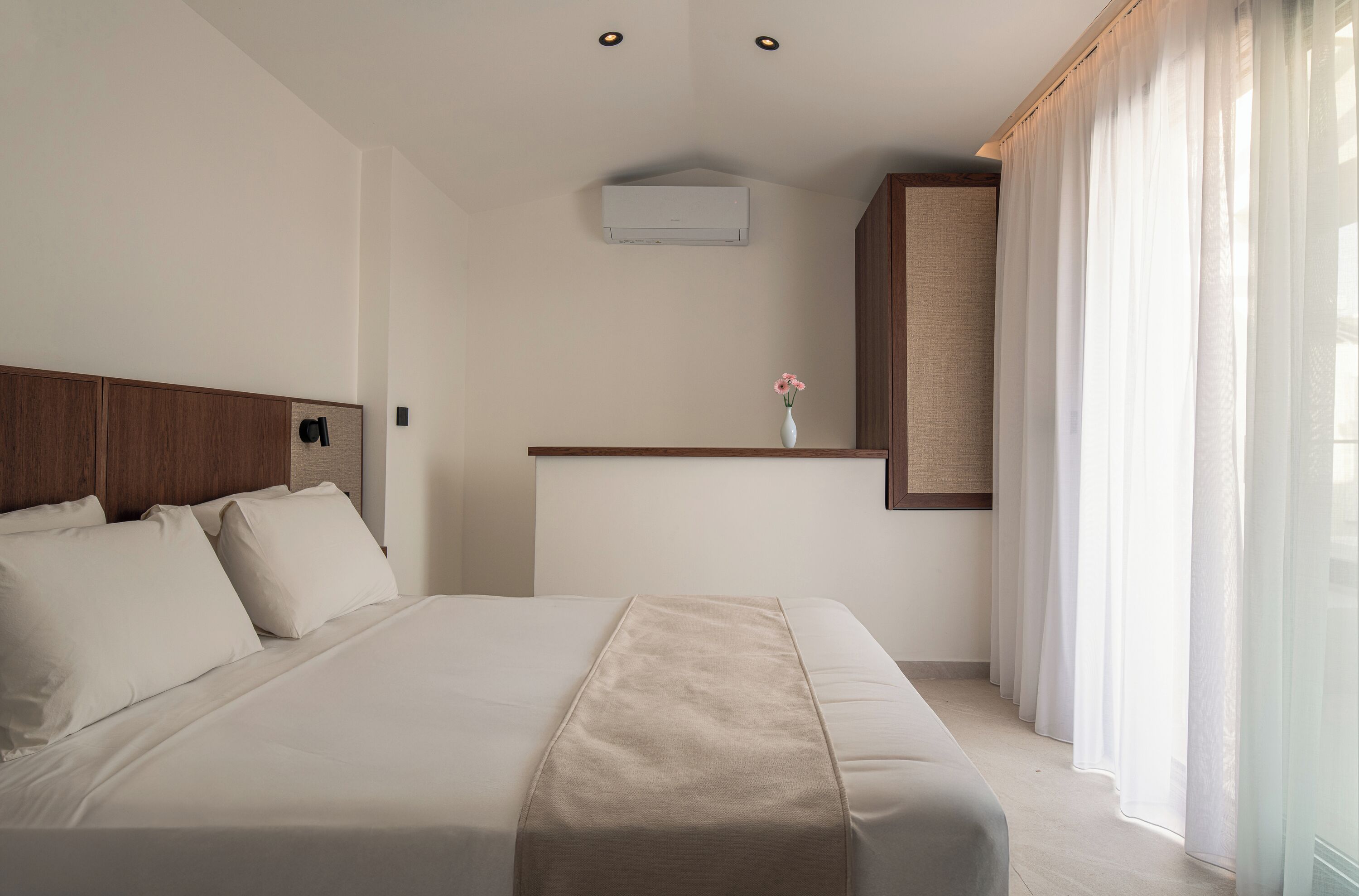 Una habitación de hotel con una cama grande, un cabecero de madera, una mesita con un florero y una ventana con cortinas.