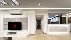 Un moderno vestíbulo de hotel con un televisor de pantalla plana mostrando el logotipo de Netflix, un mostrador de recepción y un pasillo que conduce al comedor.