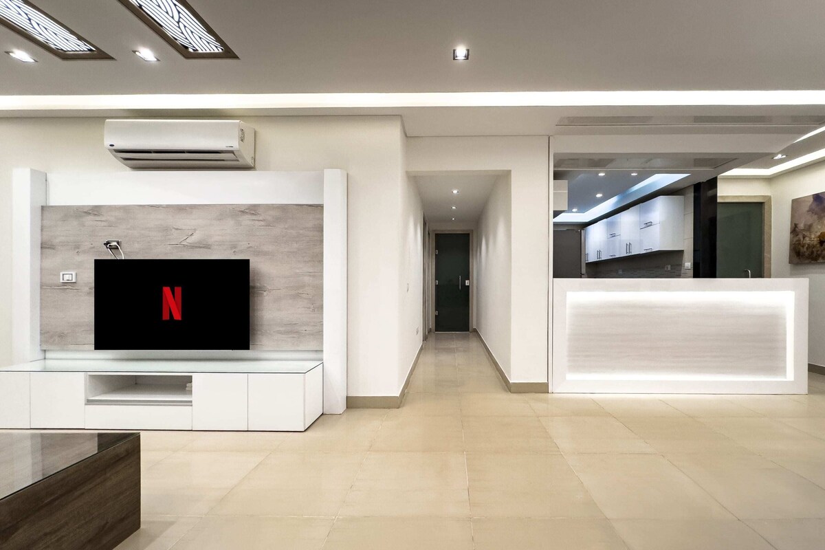 Um lobby de hotel moderno com uma TV de tela plana exibindo o logotipo da Netflix, uma recepção e um corredor que leva à área de refeições.