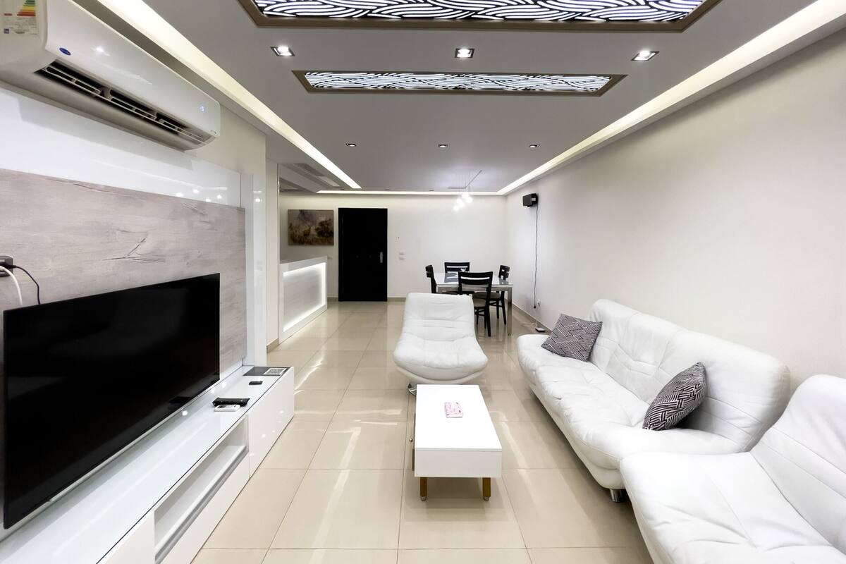 Ein modernes Wohnzimmer mit Flachbildfernseher, weißen Sofas, einem Couchtisch und Deckeneinbaustrahlern.
