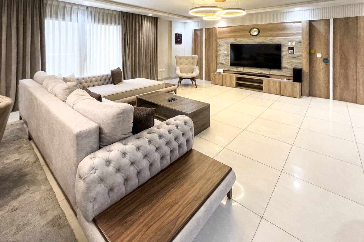 Ein modernes Wohnzimmer mit einem Ecksofa, einem gepolsterten Hocker, einem Couchtisch, einem Fernseher und einer Holzwand für Unterhaltungselektronik.