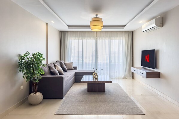 A modern living room with a sectional sofa, a coffee table, a potted plant, and a flat-screen TV mounted on the wall. - Xuru Stays I Ocean Blue - 5 Min to Airoprt (Cairo)