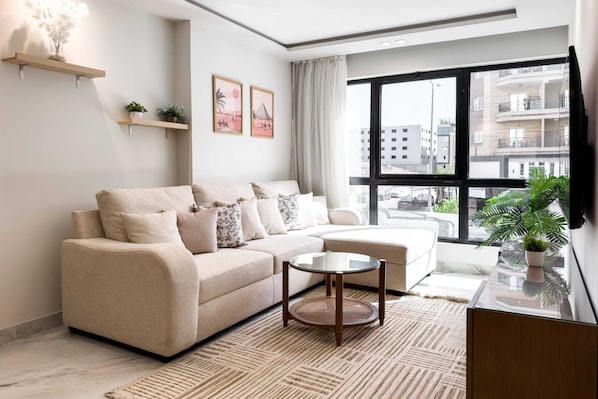 Ein modernes Wohnzimmer mit einem beige farbenen Sofa, einem Glastisch und einem großen Fenster mit Blick auf die Stadt.