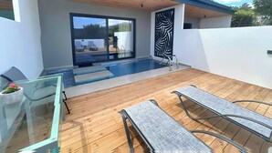 Un área de piscina exterior moderna con cerramiento de vidrio, terraza de madera y sillones de descanso.
