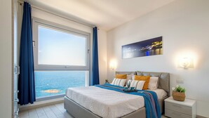 A modern bedroom with a large window, a bed with a blue and white bedspread, a nightstand with a lamp, and a wall-mounted picture. - Sardinia Re - Miramare 14 Sea View (Castelsardo)