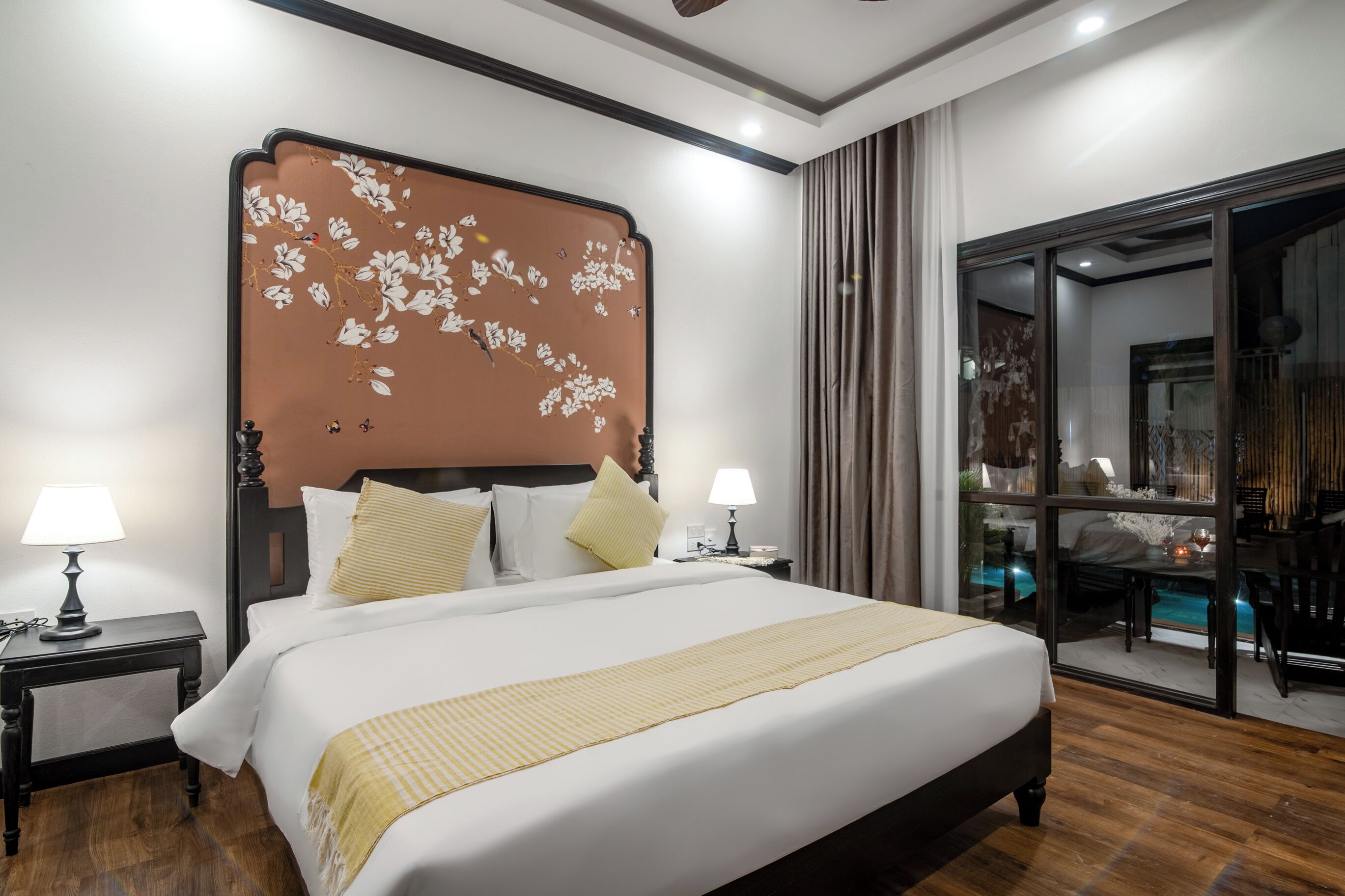 Ein Hotelzimmer mit einem großen Bett, zwei Nachttischen mit Lampen, einem Holz-Kopfteil und Blick auf einen Pool durch eine Glasdoor.