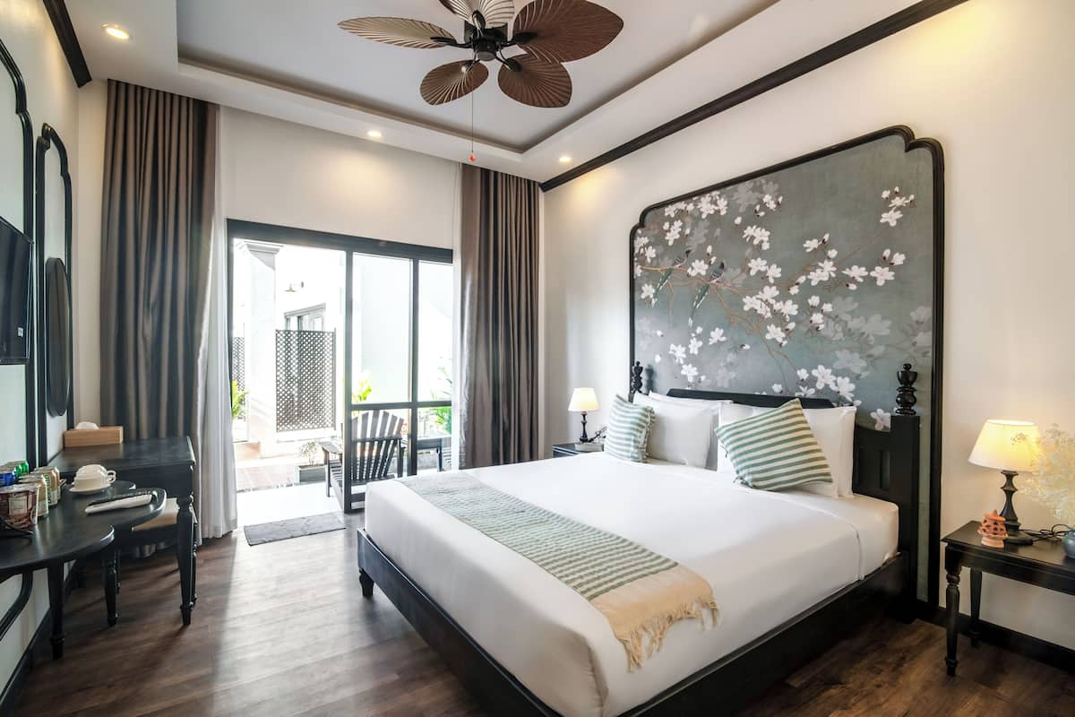 A hotel room with a large bed, a desk, a chair, a ceiling fan, and a view of the outside.
