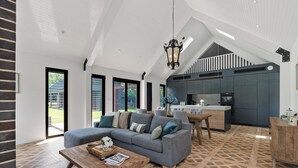 A spacious living area with a grey sofa, wooden coffee table, and a dining table. A kitchen with dark cabinets and a white countertop is visible in the background. - The Mist, Luxury Farm Retreat w/ Fireplace & Hot tub | Southern Highlands (Exeter)