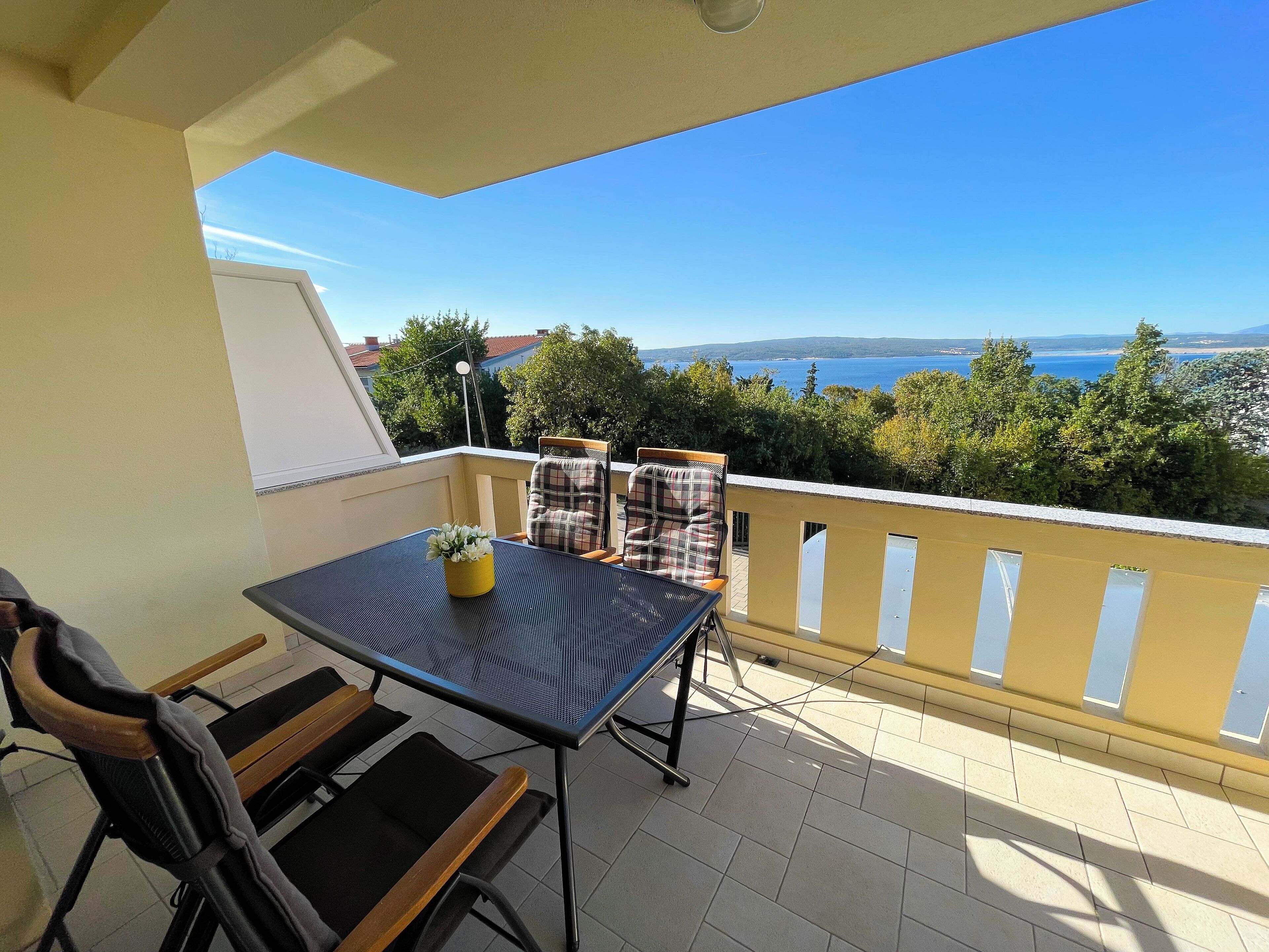 Un balcon avec une table et des chaises, un pot de fleurs et une vue sur la mer et les arbres.
