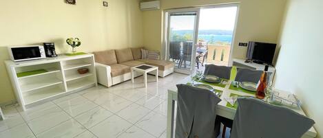 A living room with a sofa, coffee table, dining table with chairs, a TV, and a balcony with a view.
