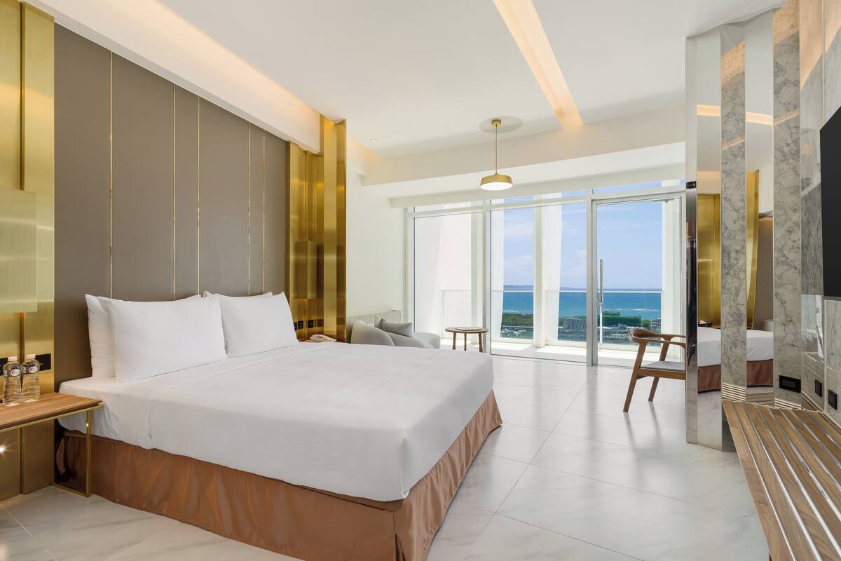 A modern hotel room with a large bed, a desk, a chair, and a view of the ocean.