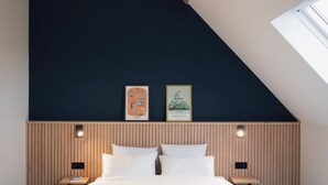 Un dormitorio con techo inclinado, una cama con ropa blanca, dos mesitas de noche fijadas a la pared y dos cuadros enmarcados.