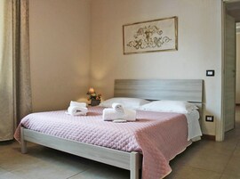 Una cama bien tendida con un edredón rosa, dos toallas enrolladas y una mesita de noche con una lámpara y flores.